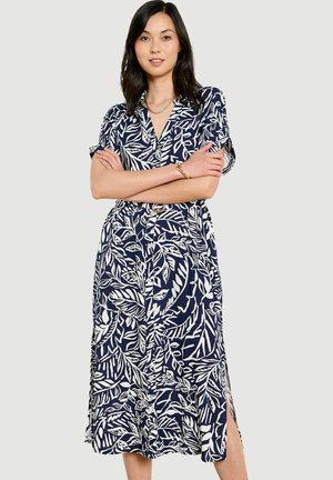 Naine käed risti, kandes tumel sinist midi kleiti valgete lehemustrite, lühikeste varrukate, külje lõhikutega ja vöökohal vööga.