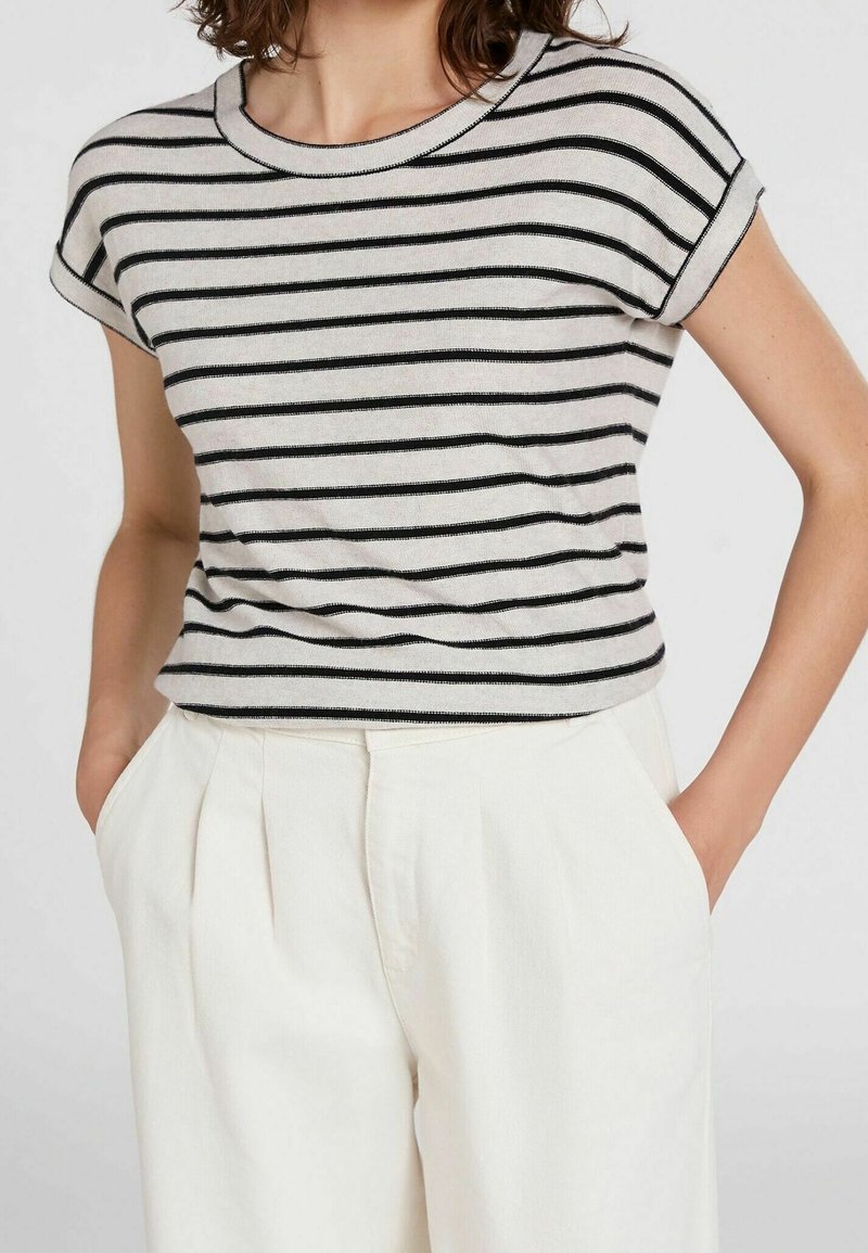 Femme portant une chemise beige à manches courtes avec des rayures horizontales noires et un pantalon blanc taille haute, les mains dans les poches.