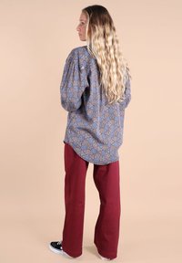 Femme aux longs cheveux blonds ondulés portant une chemise bleue à motifs, un pantalon rouge et des baskets noires, se tenant contre un fond beige, tournée vers la gauche.