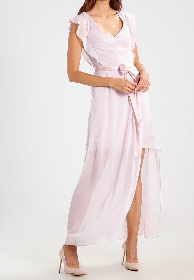 Femme portant une robe maxi sans manches rose clair avec des volants et une ceinture à la taille, associée à des chaussures à talons assorties.