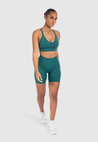 Sostén deportivo color verde azulado con tirantes delgados y frente fruncido, combinado con pantalones cortos de ciclismo de talle alto a juego. El material parece elástico y suave.