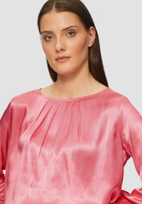 Rosa Satinbluse mit rundem Ausschnitt und gefälteltem Detail auf der Vorderseite, mit langen Ärmeln und einer glatten, glänzenden Textur.