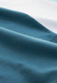Pehmeä puuvillakangas teal-värissä, jossa on sileä rakenne. Kuvassa on laskostusta hienovaraisilla varjoilla ja korostuksilla, luoden syvyyttä. Näkyvissä valkoinen yksityiskohta.