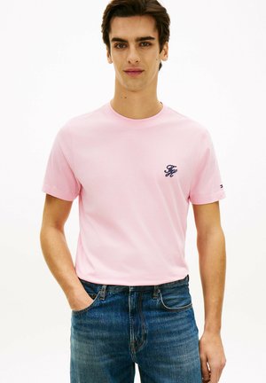 Junger Mann, der ein hellrosa T-Shirt mit einem kleinen dunklen Logo auf der Brust und blaue Jeans trägt, steht mit einer Hand in der Tasche.