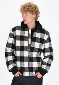 Sort-hvid ternet jakke med lynlås foran, struktureret materiale og sort krave. Har ribbede manchetter og kant.