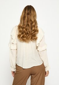 Kreemika värvi pluus, millel on õlgade ja varrukate ümber volangid, valmistatud kergest kangast, kombineeritud pruunide pükstega.