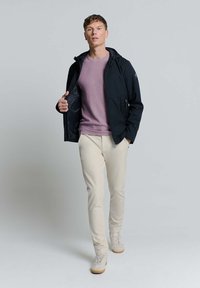 Zwarte, capuchonjack met ritszakken, gedragen over een licht paarse trui en crèmekleurige broek. Lichtgekleurde sneakers maken de outfit compleet.