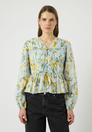 Femme aux longs cheveux bruns portant un chemisier bleu clair avec un imprimé floral jaune, trois liens à l’avant, et un jean noir taille haute, debout devant un fond blanc.