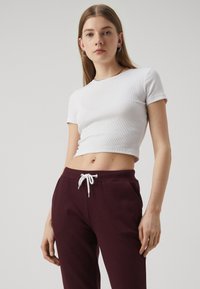 Weißes geripptes Kurzarm-Crop-Top kombiniert mit burgunderfarbenen Jogginghosen, die einen weißen Kordelzug und Seitentaschen aufweisen.