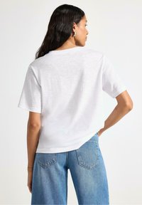 T-shirt bianco a maniche corte con vestibilità rilassata, tessuto testurizzato, scollo rotondo e orlo leggermente curvo, abbinato a jeans in denim blu chiaro.