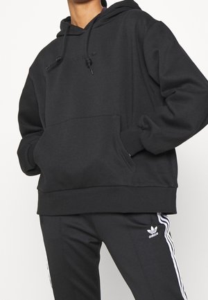 Person iført sort Adidas hættetrøje med kængurulomme og sorte Adidas træningsbukser med hvide striber og logo på siden.