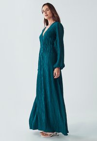 Tyrkysové maxi šaty s dlouhými rukávy vyrobené z texturované plisované látky, s hlubokým výstřihem do V a knoflíkovou ozdobou uprostřed. Nosené s bílými sandálky na pásky.