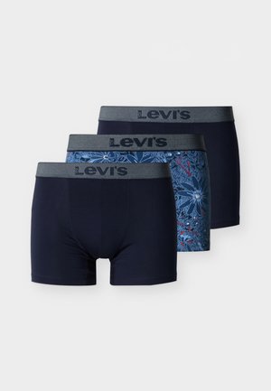 Jeune homme aux cheveux bouclés portant un boxer Levi's bleu à motif floral, debout devant un arrière-plan clair uni.