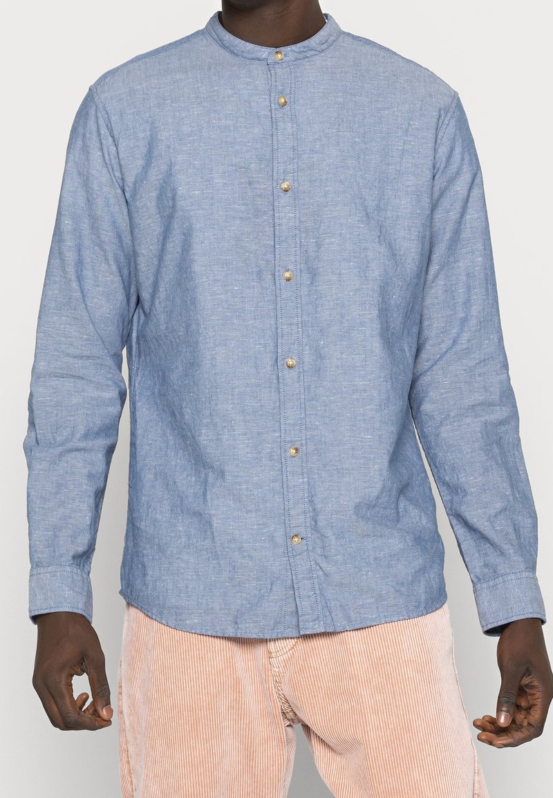 Ljusblå långärmad skjorta i texturerad tyg, med ståkrage och guldknappar framtill.