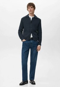 Ciemnogranatowy dzianinowy kardigan na guziki z rozłożystym kołnierzem, noszony na białej koszulce. Połączony z niebieskimi dżinsami i czarnymi butami.