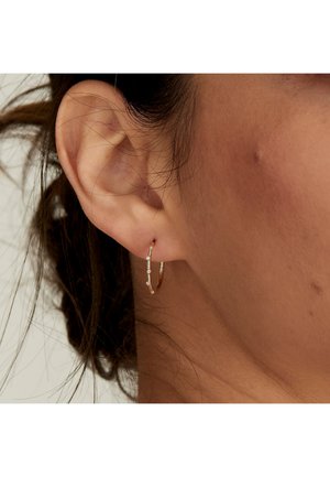 Orecchino a cerchio in oro con un design lineare e piccoli accenti trasparenti. Texture liscia con una finitura lucida, indossato su un orecchio forato.