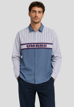 Uomo che indossa una camicia a maniche lunghe con righe verticali e logo "UMBRO" sul petto, in piedi con le mani nelle tasche.
