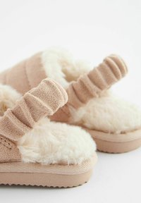 Chaussons en peluche beige avec un intérieur doux et duveteux, des bandes en tissu côtelé et des semelles en caoutchouc texturé pour plus d'adhérence.