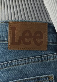 Lee logo na rjavem usnjenem znaku, prišitem na modro denim tkanino, ki ima vidne šive in teksturirano površino.