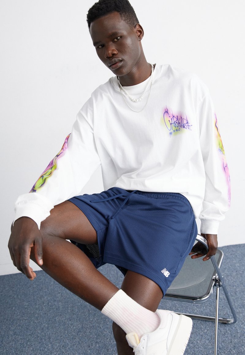 Camisa blanca de manga larga con un patrón colorido en la manga, pantalones cortos de malla azul marino, zapatos blancos y calcetines pastel, sentado en una silla de metal.