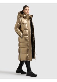 Goldfarbener, metallisch glänzender Steppmantel mit Kapuze, gesteppter Struktur, Frontreißverschluss und Seitentaschen. Trägt man über schwarzen Leggings und klobigen schwarzen Stiefeln.