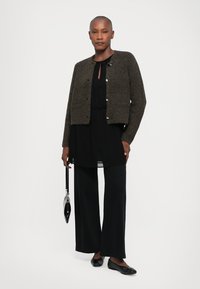 Textured olive-green cropped cardigan with silver buttons over a black flowing dress and wide-leg trousers, paired with black flats and small bag.