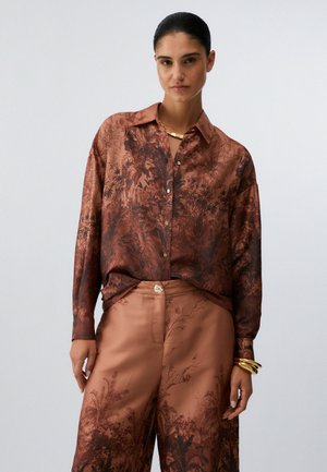 Kobieta z ciemnymi włosami spiętymi do tyłu, ubrana w brązową satynową koszulę i pasujące szerokie spodnie z ciemnymi botanicznymi wzorami oraz złotą biżuterią.