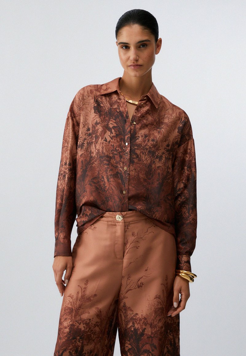 Femme aux cheveux foncés attachés en arrière, portant une chemise en satin marron et un pantalon ample assorti avec des motifs botaniques foncés, ainsi que des bijoux dorés.