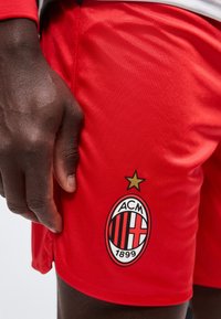 Κοντινό πλάνο του χεριού ενός ατόμου που ακουμπά κοντά σε κόκκινα ποδοσφαιρικά σορτς της AC Milan με το έμβλημα του συλλόγου και ένα χρυσό αστέρι πάνω από αυτό.