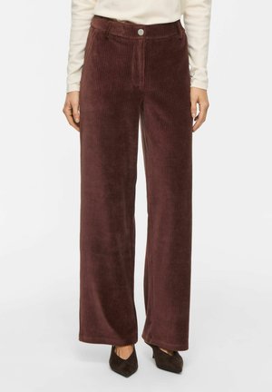 Pantalon large en velours côtelé marron porté par une personne debout, associé à des chaussures pointues marron foncé et un haut crème à manches longues.