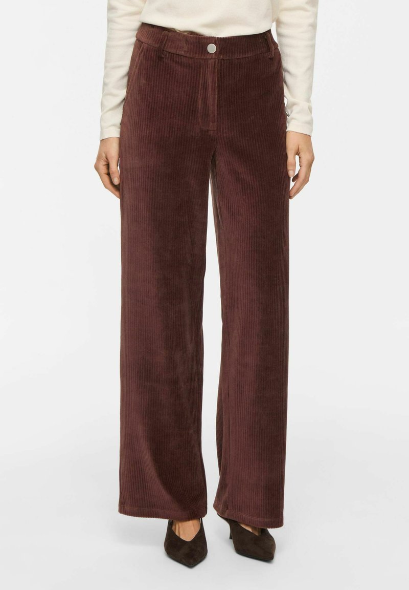 Pantalon large en velours côtelé marron porté par une personne debout, associé à des chaussures pointues marron foncé et un haut crème à manches longues.