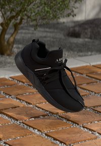 Black athletic shoe with a textured mesh upper, featuring a sleek design, laces, and subtle branding accent. Sole designed for traction.