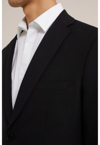 Blazer noir avec une coupe ajustée, un revers en pointe et une poche plaquée, associé à une chemise habillée blanche visible au niveau du col.