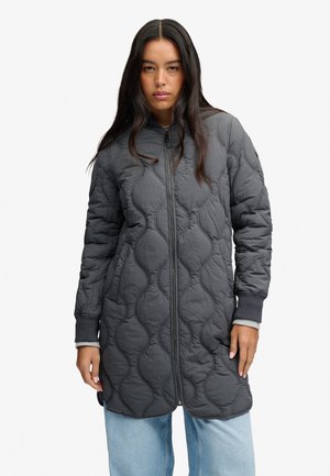 Jeune femme portant une longue veste matelassée gris foncé avec une fermeture éclair à l'avant et un jean bleu clair, debout devant un fond blanc uni.