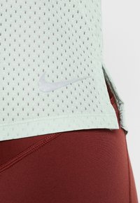 Top atlético de malla transpirable en verde claro con pequeños orificios, con un logo de Nike plateado, combinado con leggings ajustados de color rojo profundo.