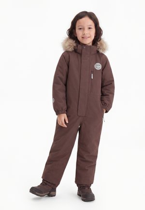 Criança sorridente vestindo um macacão de inverno isolado castanho com capuz forrado a pelo e botas de inverno castanhas, a posar sobre um fundo branco.