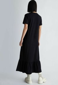 Musta maksimekko lyhyillä hihoilla ja röyhelöhelmalla. Mallilla on päällään kermanväriset, paksupohjaiset saappaat, joissa on rei'itetty kuvio.