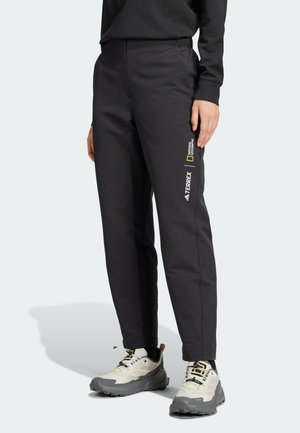 Personne portant un pantalon de sport noir avec les logos National Geographic et Terrex sur la jambe droite, associé à des chaussures de randonnée blanches et grises.