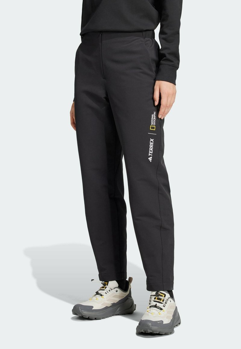 Personne portant un pantalon de sport noir avec les logos National Geographic et Terrex sur la jambe droite, associé à des chaussures de randonnée blanches et grises.