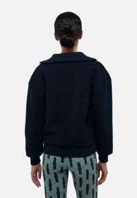 Veste texturée navy avec un large col, coupe oversize, poignets côtelés et ourlet. Associée à un legging à motifs avec des lignes bleues et vertes.