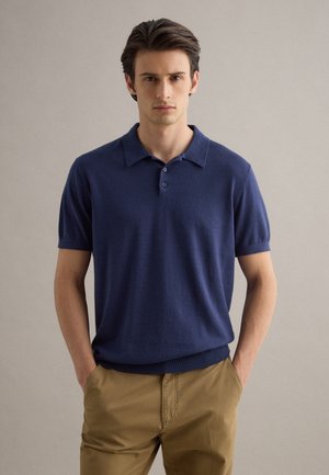 Giovane uomo in piedi con le mani nelle tasche, indossa una polo a maniche corte blu navy e pantaloni marrone chiaro su uno sfondo semplice.