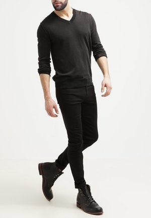 Schwarzer V-Ausschnitt-Pullover mit langen Ärmeln, kombiniert mit enganliegenden schwarzen Hosen und schwarzen Schnür-Ankle-Boots mit Akzentnähten.