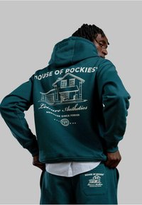 Felpa teal con design grafico bianco di una casa, testo "House of Pockies" e estetica da tempo libero. Materiale in cotone con vestibilità rilassata.