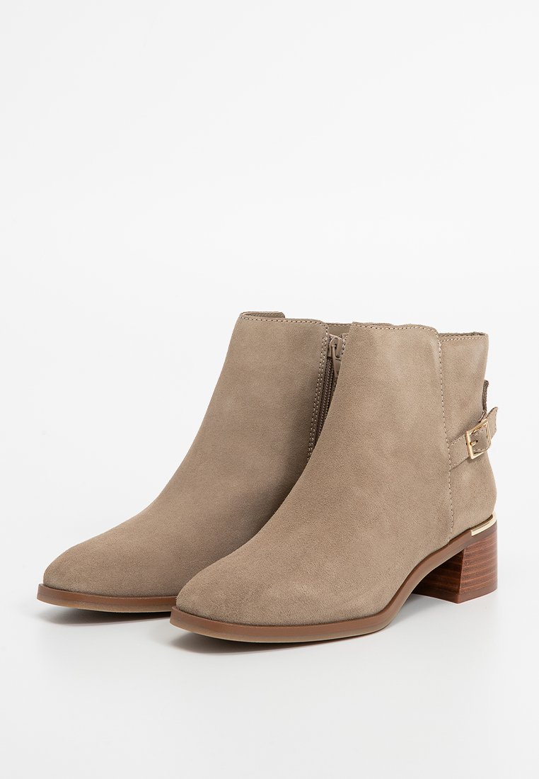Bottines en daim beige avec un bout rond, une fermeture éclair sur le côté et un accent en boucle dorée. Le talon en bois surélevé ajoute de la stabilité.