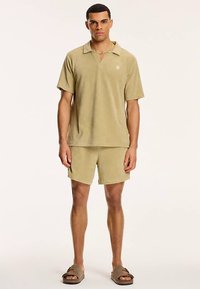Ensemble chemise et short en tissu éponge kaki avec col, manches courtes et détail logo. Le modèle porte des sandales marron.