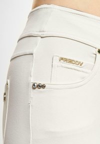 Hvite stretchjeans med gull "FREDDY"-logo og dekorative metallstuds på baklommen. Har en tettsittende design og en glatt tekstur.