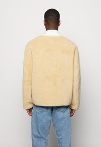 Giacca oversized in pile beige con una morbida texture, spalle arrotondate e orlo corto, abbinata a jeans in denim azzurro chiaro.