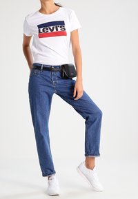 Camiseta blanca con logo de Levi's en rojo y azul, jeans azules, riñonera negra, zapatillas blancas, corte relajado, material de algodón, diseño casual.