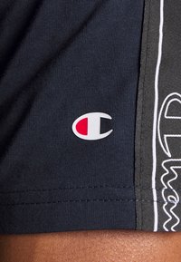 Logo značky Champion na černé látce vedle bočního panelu v bílé a černé barvě se stylizovaným textem na sportovním oblečení.