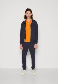 Marineblå hoodie med lynlås, orange polo-shirt under, marineblå bukser og hvide sneakers. Glat tekstur, afslappet design og kontrasterende farver.